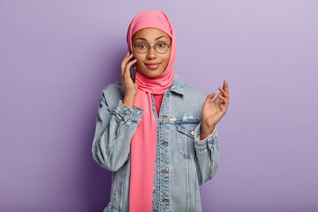 Taille-up shot van moslimvrouw geniet van smartphone-gesprek, als geavanceerde gebruiker van modern apparaat, draagt roze hijab en spijkerjasje, maakt gebruik van openbare internetverbinding, geïsoleerd op paarse muur