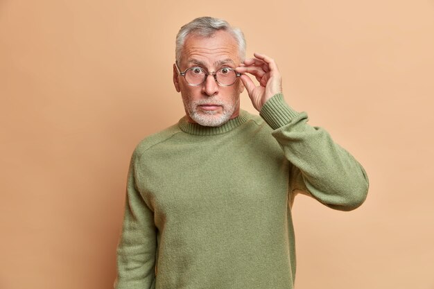 Taille-up shot van verbijsterde senior man staart door bril reageert op onverwacht nieuws wordt geschokt poses verstoord tegen beige muur draagt vrijetijdskleding