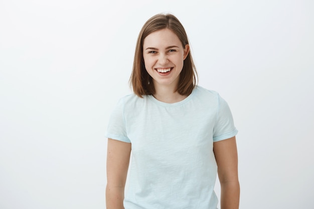 Taille-up shot van vrolijk vermaakt en geamuseerd goed uitziende gewone brunette meisje in trendy t-shirt vrolijk lachen en staren met grappige grappige praat met vrienden over grijze muur