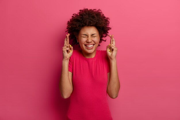 Taille-up shot van vrolijke jonge Afro-Amerikaanse vrouw kruist vingers voor geluk, verwacht dat er iets goeds gebeurt, wacht op een groot fortuin, draagt een roze t-shirt, stelt zich voor dat dromen uitkomen