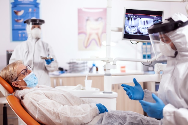Tandarts die veiligheidsuitrusting draagt tegen coronavirus die over tandenbehandeling spreekt. Oudere vrouw in beschermend uniform tijdens medisch onderzoek in tandheelkundige kliniek.