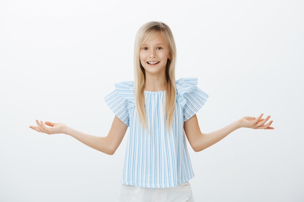 Gratis foto te veel keuzes voor één kind. portret van clueless verward aantrekkelijk jong blond meisje in blauwe blouse, handpalm spreiden en wegen of schouders ophalen, ondervraagd en zich niet bewust over grijze muur