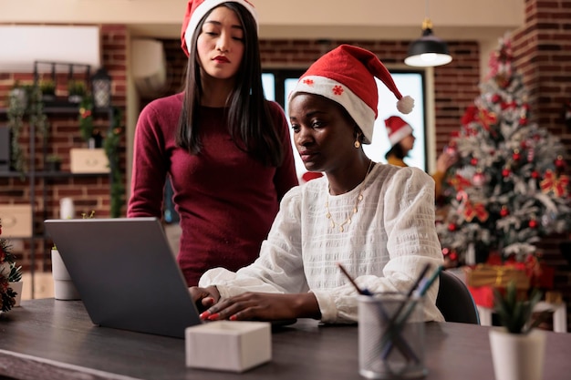 Team van collega's die aan zaken werken op de werkplek, versierd met kerstverlichting, boom- en vakantieornamenten. Laptop gebruiken in feestelijk bedrijfskantoor met seizoensgebonden kerstversieringen.