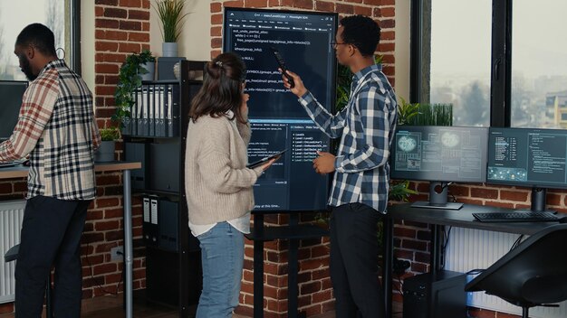 Team van databasebeheerders die de broncode analyseren op tv op het muurscherm en fouten vergelijken met behulp van digitale tablet in drukke serverruimte. Twee cloudprogrammeurs debuggen algoritme in software-innovatiekantoor.