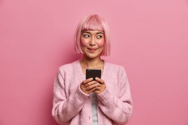 Technologie, communicatie en levensstijlconcept. Blij dromerige vrouw met geverfd roze haar, houdt mobiele telefoon vast, gebruikt moderne app om berichten te sturen met vrienden,