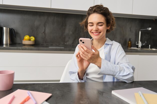 Technologie en levensstijl jonge vrouw zit thuis gebruikt smartphone in haar keuken en glimlacht