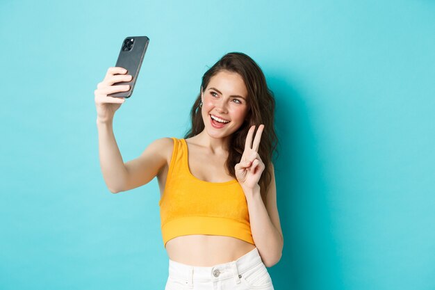 Technologie en lifestyle concept. Stijlvolle jonge vrouw die selfie in haar zomerkleren neemt, v-teken toont op smartphonecamera, staande over blauwe achtergrond