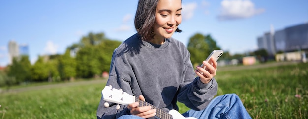 Gratis foto technologie en muziek glimlachend aziatisch hipster meisje dat in het park zit met een smartphone en een ukulele vasthoudt