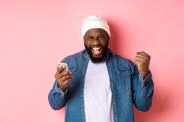 Technologie en online winkelconcept. Gelukkige zwarte man verheugt zich, wint in app, houdt smartphone vast en schreeuwt ja, staande over roze achtergrond