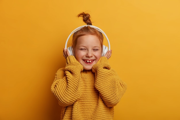 Technologie, kinderen, muziekconcept. Vrij glimlachend klein kind met rood haar draagt een stereohoofdtelefoon, geniet van puur geluid en luistert naar favoriete liedje, giechelt vrolijk, draagt een gebreide trui