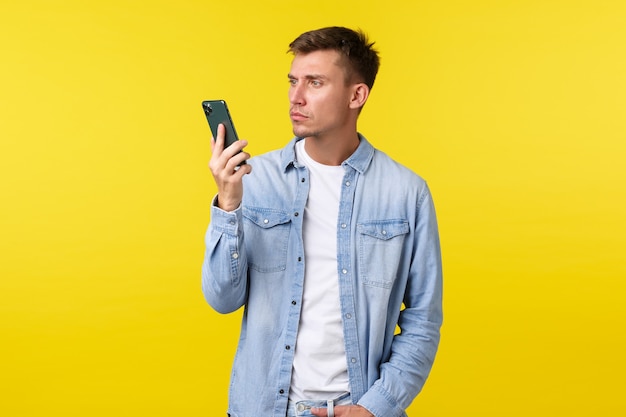 Technologie, lifestyle en reclame concept. Geërgerde en gefrustreerde jonge man verbaasd over plotseling beëindigd gesprek, kijkend naar geïrriteerde mobiele telefoon, staande over gele achtergrond.