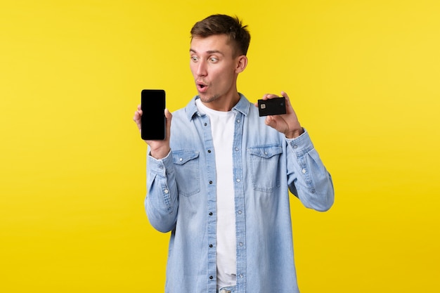 Technologie, lifestyle en reclame concept. onder de indruk en verbaasde blonde man die wauw zegt terwijl hij naar het scherm van de mobiele telefoon kijkt, smartphone-display en creditcard toont, bestelling plaatst.