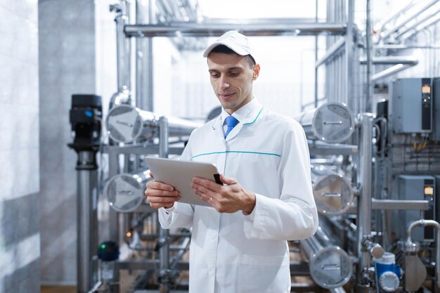 Technoloog in een witte jas maakt de nodige vermeldingen in de tablet is in de fabriek Een man met een digitale tablet in uniform in het productiewinkelinterieur van de productieafdeling op de achtergrond