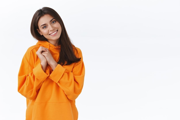 Teder, vriendelijk ogend vrouwelijk brunette meisje in oranje hoodie, kantelend hoofd en glimlachend aangenaam camera