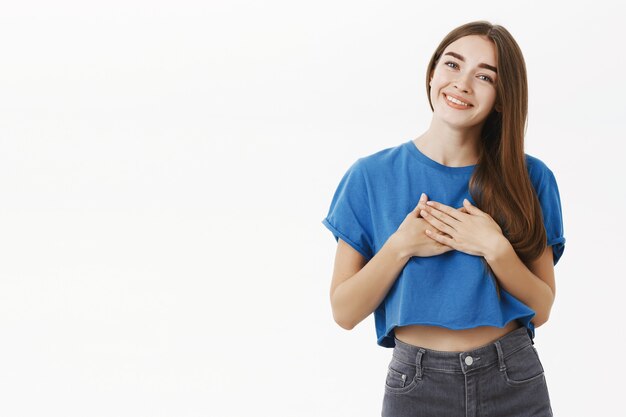 Tedere charmante vrouw met bruin haar in blauw t-shirt met handpalmen op hart in dankbaar gebaar kantelend hoofd gefascineerd en aangeraakt met schattige glimlach staren