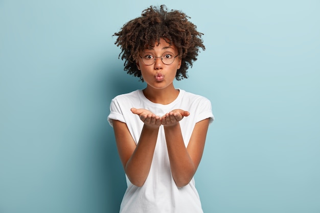 Gratis foto tedere gevoelens. mooie krullende vrouw met afro-kapsel, vouwt de lippen en stuurt luchtkus, geeft mwah, strekt de handpalmen uit, draagt een transparante bril voor zichtcorrectie, geïsoleerd over blauwe muur