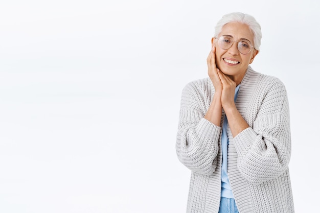 Tedere mooie senior vrouw met grijs gekamd haar in bril op sterkte, gezellig vest, ontroerende wang en glimlachend hartverwarmend