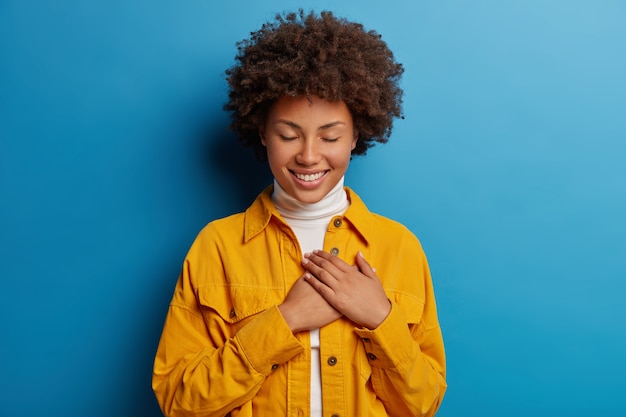 Tedere vrouwelijke vrouw houdt de handen op het hart, heeft een dankbare blik, waardeert moeite, heeft de ogen dicht, draagt een geel shirt, geïsoleerd op blauwe achtergrond