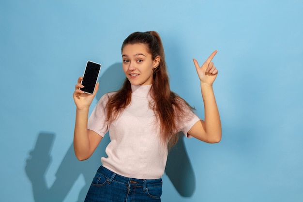 Telefoonscherm wordt weergegeven, met de punt omhoog. Het portret van het Kaukasische tienermeisje op blauwe achtergrond. Prachtig model in vrijetijdskleding. Concept van menselijke emoties, gezichtsuitdrukking, verkoop, advertentie. Copyspace. Ziet er blij uit.