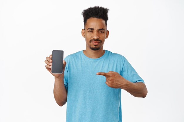 Teleurgesteld Afro-Amerikaanse man wijzende vinger naar smartphone scherm met afkeer, wenkbrauwen fronsen ontevreden, afkeer interface, slechte toepassing, staande in t-shirt tegen witte achtergrond