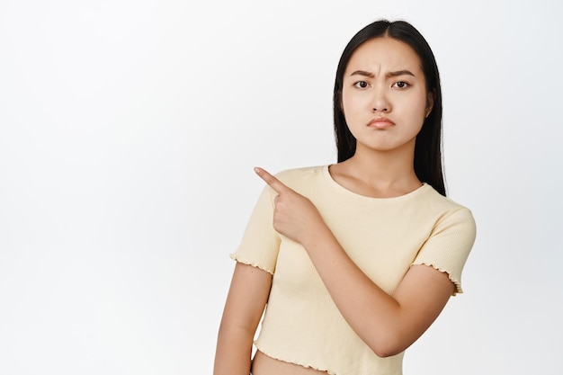 Teleurgesteld jong Aziatisch meisje kijkt sceptisch naar de vinger in de linkerbovenhoek en toont advertentie witte achtergrond