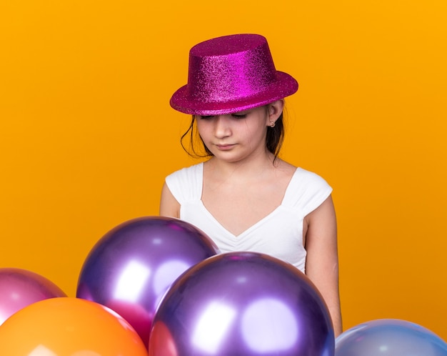 teleurgesteld jong kaukasisch meisje met paarse feestmuts kijkend naar heliumballonnen geïsoleerd op een oranje muur met kopieerruimte