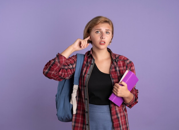 Teleurgesteld jong slavisch studentenmeisje dat rugzak draagt, houdt boek vast en notitieboekje sluit oor met vinger