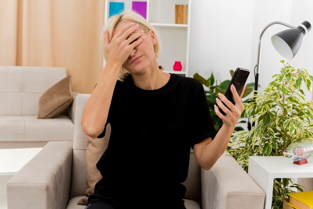 Teleurgesteld mooie blonde Russische vrouw zit op fauteuil hand op gezicht houden telefoon in de woonkamer