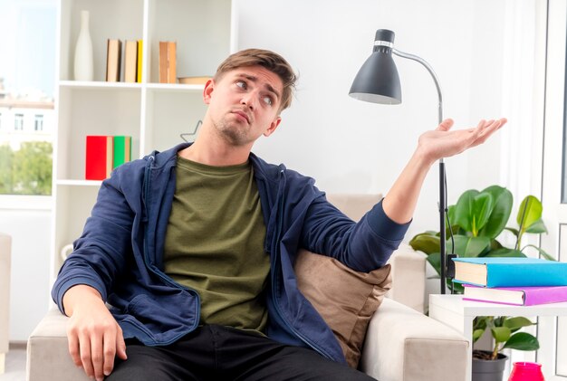 Teleurgestelde jonge blonde knappe man zit op fauteuil looing en wijzend naar de zijkant met de hand