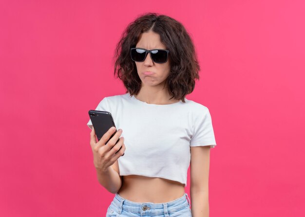 Teleurgestelde jonge mooie vrouw die zonnebril draagt en mobiele telefoon op geïsoleerde roze muur met exemplaarruimte houdt