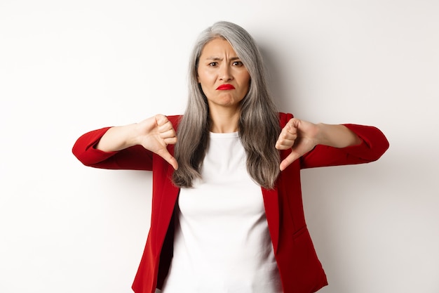 Teleurgestelde oudere vrouw in rode blazer met thumbs-down, grimassen boos, afkeer en afkeuren, staande tegen een witte achtergrond.