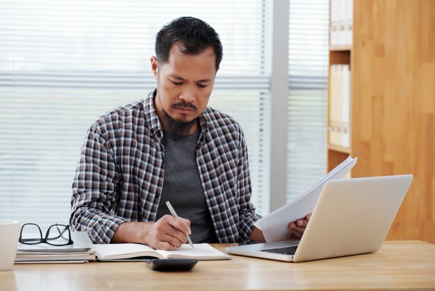 Terloops geklede Aziatische man die in bureau werkt, in notitieboekje schrijft en documenten houdt