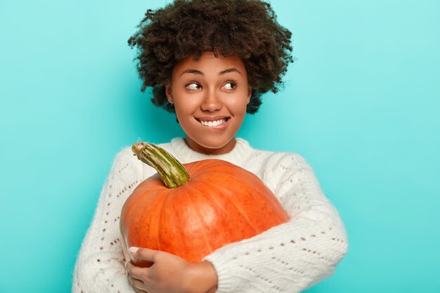 Tevreden Afro-meisje omarmt grote oranje pompoen, bijt lippen, draagt gebreide witte trui, heeft herfststemming, kijkt opzij, geïsoleerd op blauwe achtergrond.