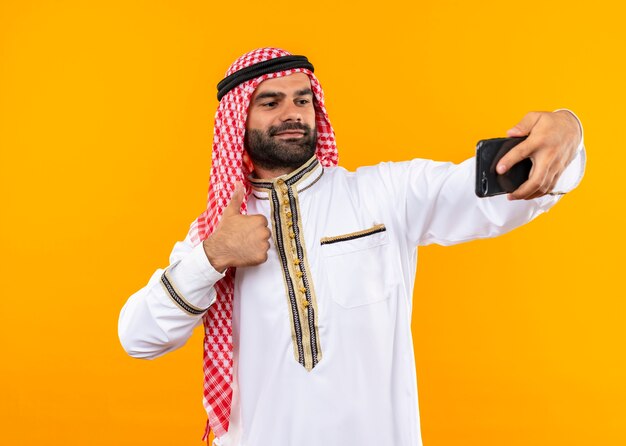 Tevreden Arabische zakenman in traditionele slijtage die selfie met zijn smartphone neemt die zelfverzekerd over oranje muur kijkt