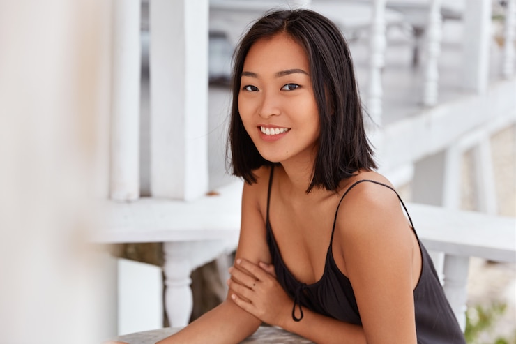 Gratis Foto tevreden aziatische vrouw met brede glimlach heeft kapsel afgeknipt, terloops gekleed, zit aan café tafel`` geniet van recreatietijd. mooie japanse vrouw rust alleen in restaurant