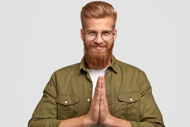 Tevreden bebaarde man met geperste handpalmen, heeft een vrolijke uitdrukking, gelooft in iets goeds, gekleed in een modieuze outfit, geïsoleerd op een witte muur. mensen en lichaamstaal concept