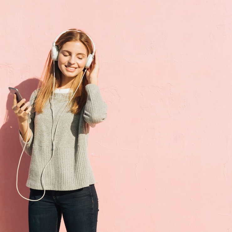 Gratis Foto tevreden blonde jonge vrouw die van de muziek op mobiele telefoon genieten door hoofdtelefoon tegen roze achtergrond