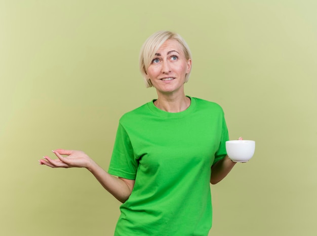 Tevreden blonde slavische vrouw die op middelbare leeftijd kop thee bekijkt die kant bekijkt die lege hand toont die op olijfgroene muur wordt geïsoleerd