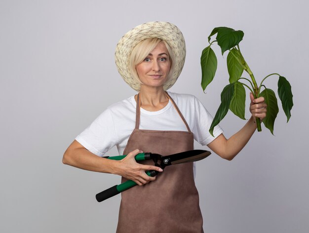 Tevreden blonde tuinmanvrouw van middelbare leeftijd in uniform met hoed met plant en heggenschaar geïsoleerd op een witte muur