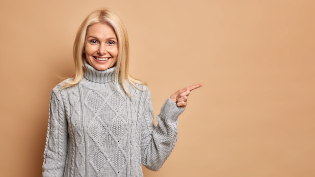 tevreden blonde vrouw van middelbare leeftijd met rimpels draagt warme grijze trui, wijzend op kopie ruimte