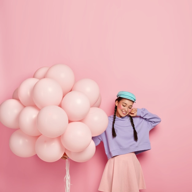 Tevreden brunette vrouw met twee vlechten, stijlvolle pet, paarse losse sweatshirt en roze rok, houdt helium ballonnen