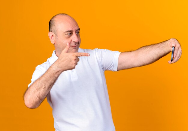 Tevreden casual volwassen man neemt een selfie en wijst naar telefoon geïsoleerd op gele muur