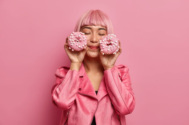 Tevreden charmante vrouw met roze haar en pony, sluit de ogen, stelt zich een aangename smaak van donuts voor, gekleed in een roze jasje