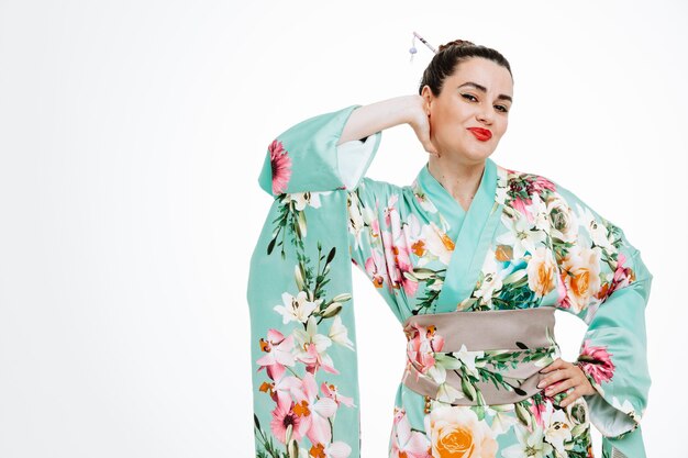 Tevreden en gelukkige vrouw in traditionele Japanse kimono glimlachend met hand op haar hoofd en andere hand op heup op wit