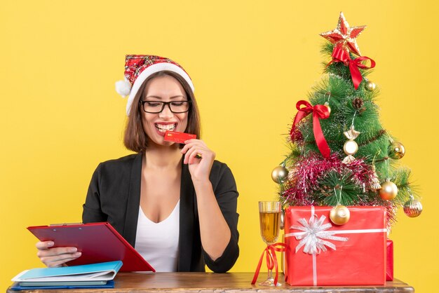 Tevreden gelukkig lachend charmante dame in pak met kerstman hoed en bril met bankkaart in het kantoor op geel geïsoleerd