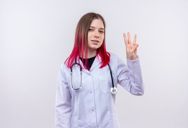 Tevreden jong artsenmeisje dat stethoscoop medisch kleed draagt dat drie op geïsoleerde witte achtergrond toont