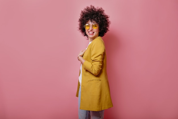 Tevreden jong glimlachend meisje die met krullen geel jasje dragen die voorzijde over roze muur bekijken