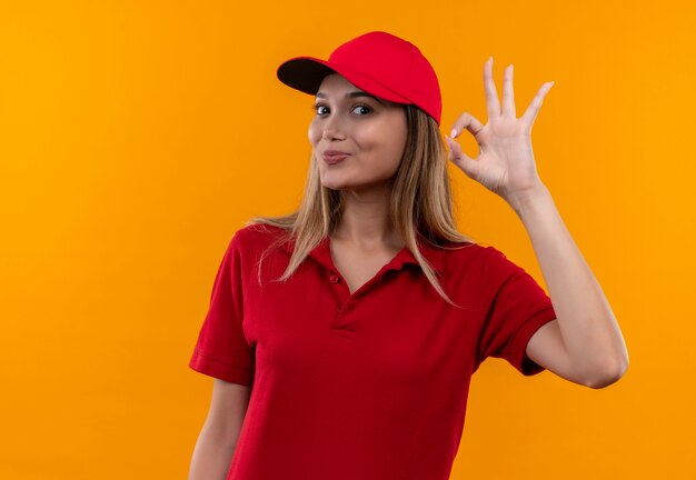 Tevreden jong leveringsmeisje die rood uniform en GLB dragen die okgebaar tonen dat op oranje achtergrond wordt geïsoleerd