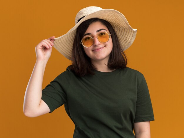 Tevreden jong mooi Kaukasisch meisje in zonnebril en met strandhoed geïsoleerd op een oranje muur met kopieerruimte