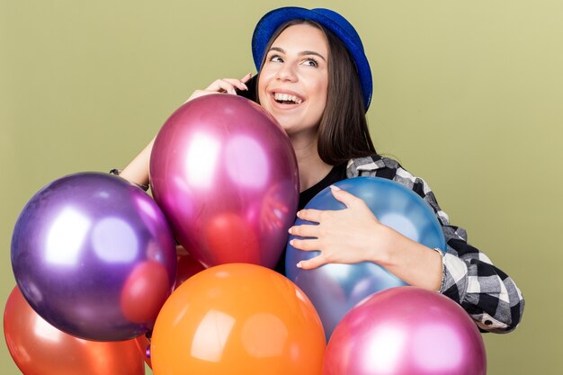 Tevreden jong mooi meisje met blauwe hoed die achter ballonnen staat, spreekt aan de telefoon